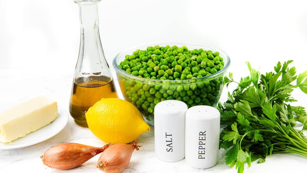 A bowl of green peas, fresh parsley, a lemon, two shallots for extra flavor, creamy butter, olive oil, and salt and pepper shakers are arranged on a white surface—perfect ingredients for a classic Buttered Peas Recipe.