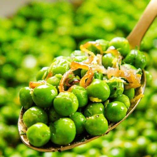A close-up of a spoon holding bright green buttered peas mixed with caramelized shallots, with more peas blurred in the background.