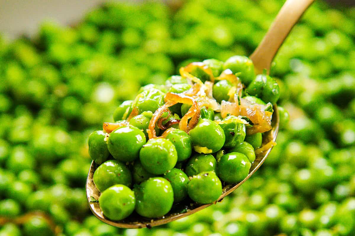 A close-up of a spoon holding bright green buttered peas mixed with caramelized shallots, with more peas blurred in the background.
