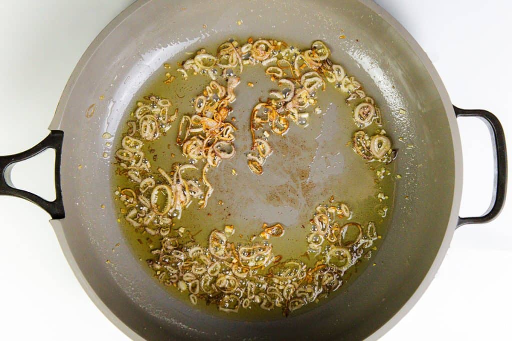 Sliced shallots being sautéed in oil in a large, round, gray pan with black handles. The shallots are turning golden brown and are spread out in the hot oil—perfect for adding to your favorite Buttered Peas Recipe.