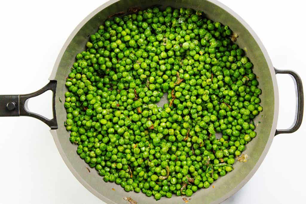 A top-down view of a frying pan filled with sautéed green peas and bits of caramelized shallots, against a white background—a simple yet elegant Buttered Peas recipe.