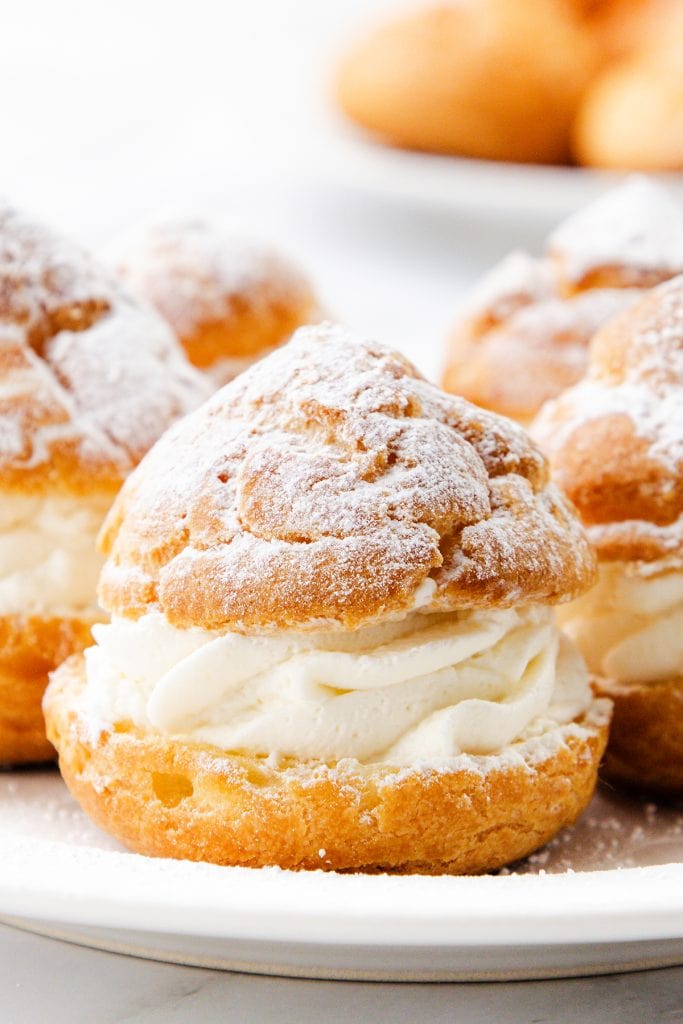 Close-up of cream puffs filled with whipped cream and dusted with powdered sugar, arranged on a white plate with more cream puffs blurred in the background.