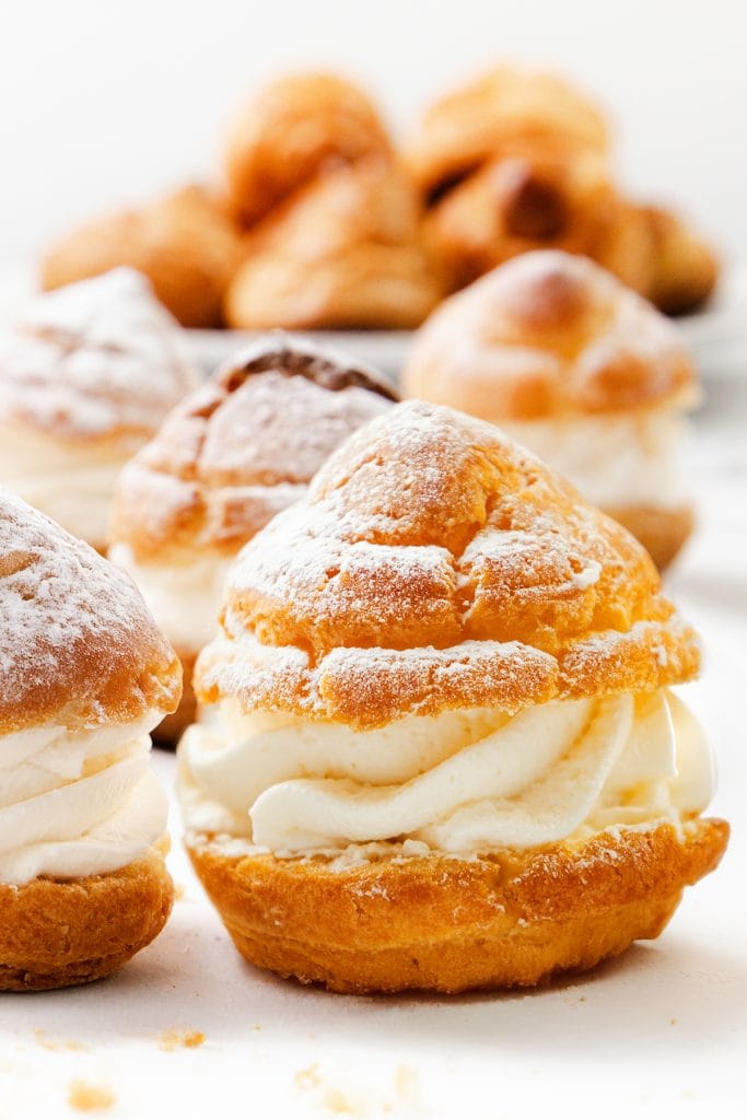 A close-up of cream puffs filled with whipped cream and dusted with powdered sugar, with more cream puffs blurred in the background.