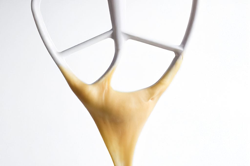 A close-up of a soft dough texture, and thick Choux Pastry falling from a paddle attachment in a soft V shape downward against a plain white background.