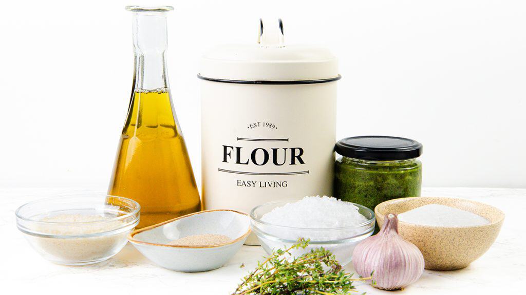 A collection of cooking ingredients including olive oil, a flour canister, Parmesan, a jar of pesto, garlic, fresh thyme, and bowls of breadcrumbs, salt, and sugar—all arranged on a white surface.