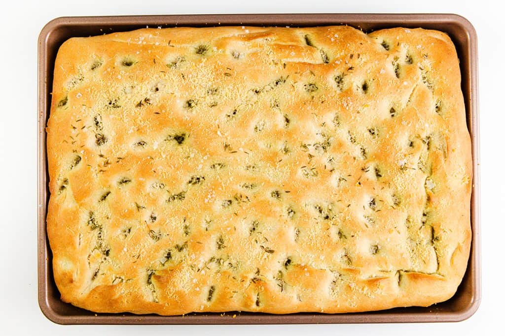 A golden-brown garlic focaccia bread with dimples, a sprinkling of herbs, coarse salt, and a touch of Parmesan, baked in a rectangular metal pan.