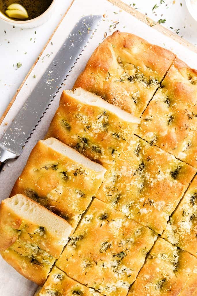 A golden, sliced Garlic Focaccia bread topped with herbs and grated Parmesan sits on parchment paper next to a serrated knife and a small bowl of olive oil.