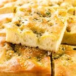 Golden, fluffy slices of garlic focaccia bread topped with herbs, Parmesan, and coarse salt are stacked on parchment paper, with a blurred bowl in the background.