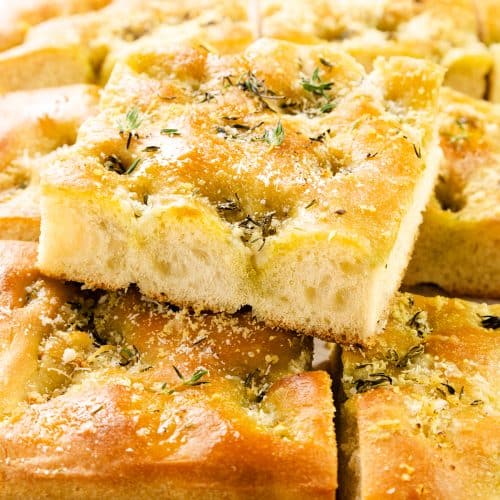 Golden, fluffy slices of garlic focaccia bread topped with herbs, Parmesan, and coarse salt are stacked on parchment paper, with a blurred bowl in the background.