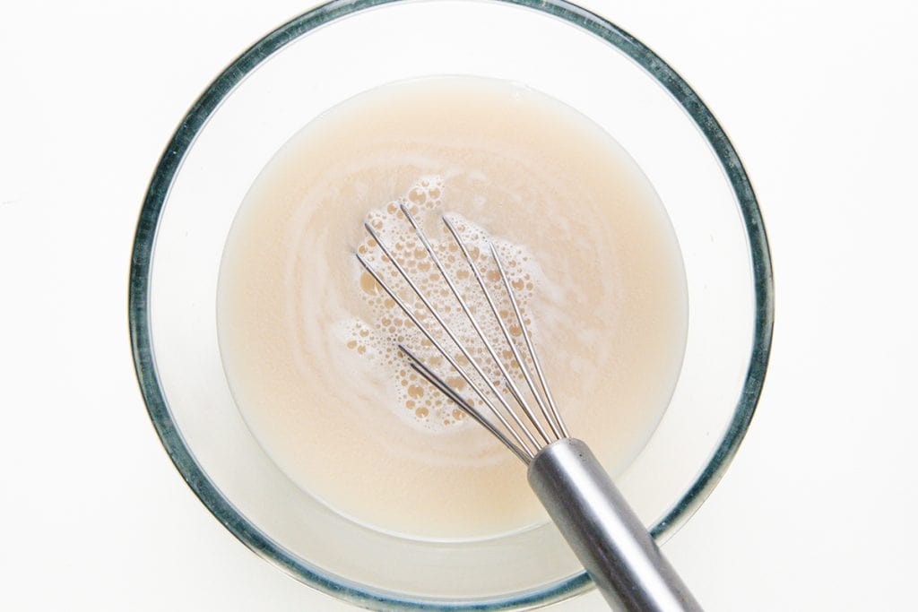 A metal whisk sits in a glass bowl filled with a bubbly, light beige liquid, likely a mixture of yeast and water ready to become garlic focaccia, on a white background.