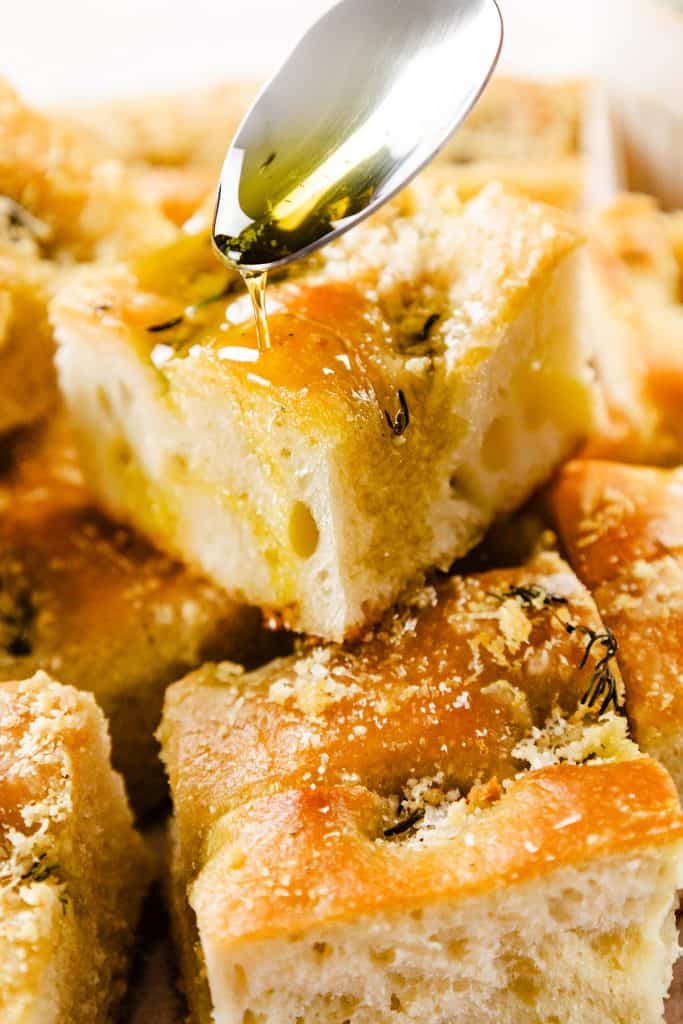 A close-up of golden, fluffy garlic focaccia bread squares being drizzled with olive oil from a spoon, highlighting their airy texture and savory topping.