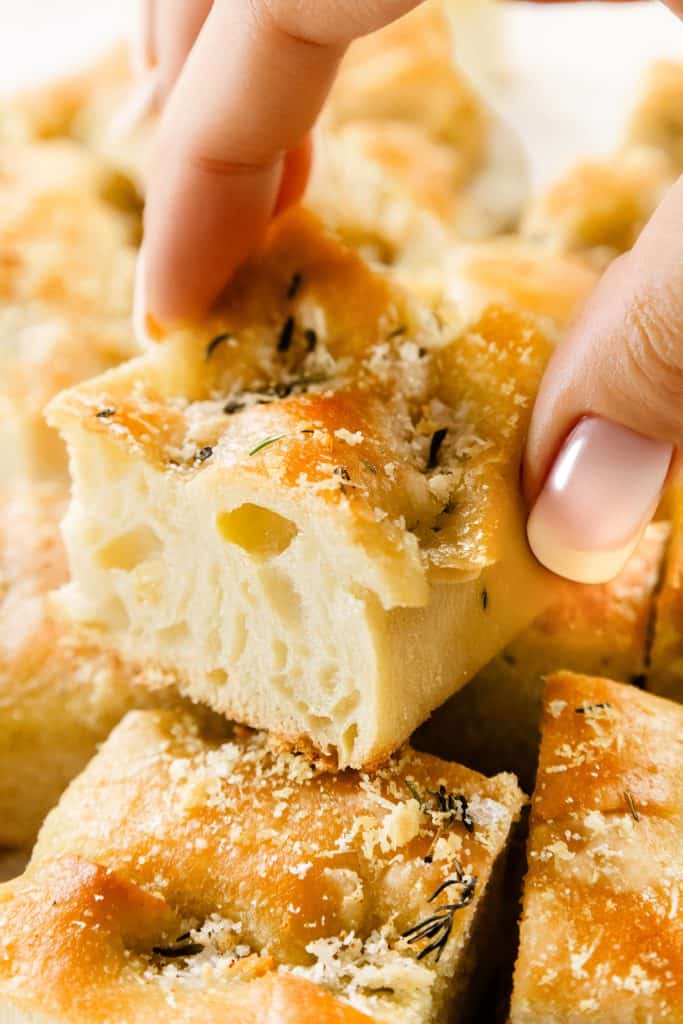 A close-up of a hand with manicured nails picking up a square piece of golden, fluffy garlic focaccia bread topped with herbs and grated Parmesan from a pile of similar pieces.