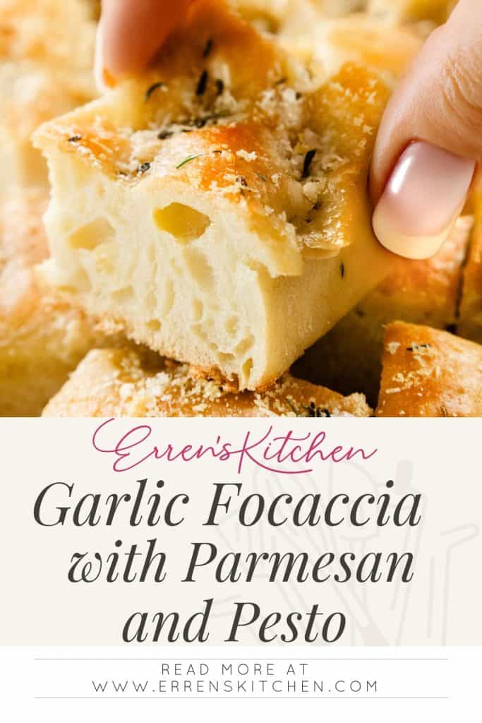 A close-up of a hand holding a piece of garlic focaccia topped with Parmesan and herbs, plus a drizzle of pesto, with more slices in the background. Text below reads: "Garlic Focaccia with Parmesan and Pesto.