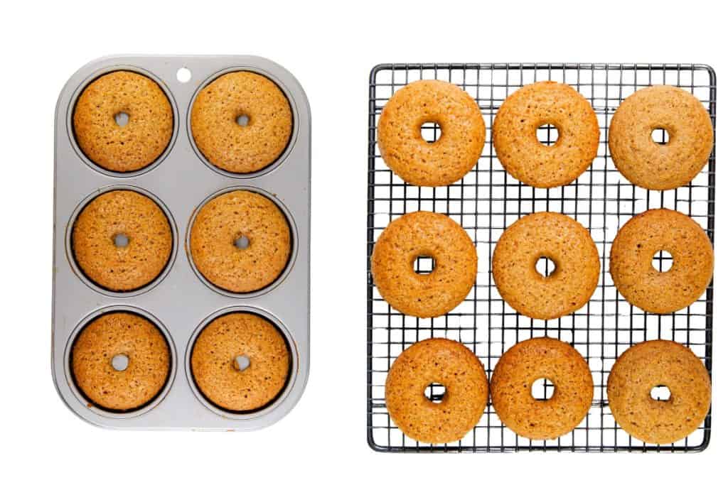 A metal tray with six baked gingerbread donuts sits next to a cooling rack holding twelve more donuts, all with holes in the center, on a white background.