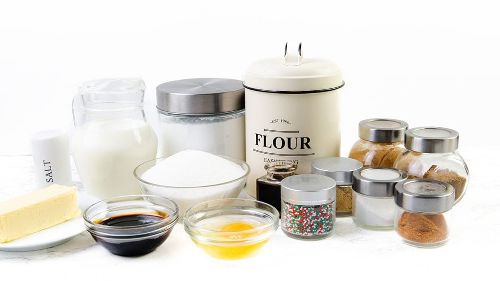 An assortment of baking ingredients on a white surface, including flour, sugar, milk, butter, vanilla, honey, salt, spices in jars—perfect for making gingerbread donuts—and colorful sprinkles in glass bowls and containers.