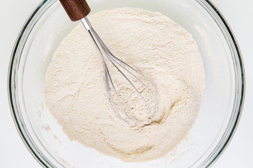 A metal whisk with a wooden handle rests in a glass bowl filled with flour, ready for mixing up gingerbread donuts, viewed from above.