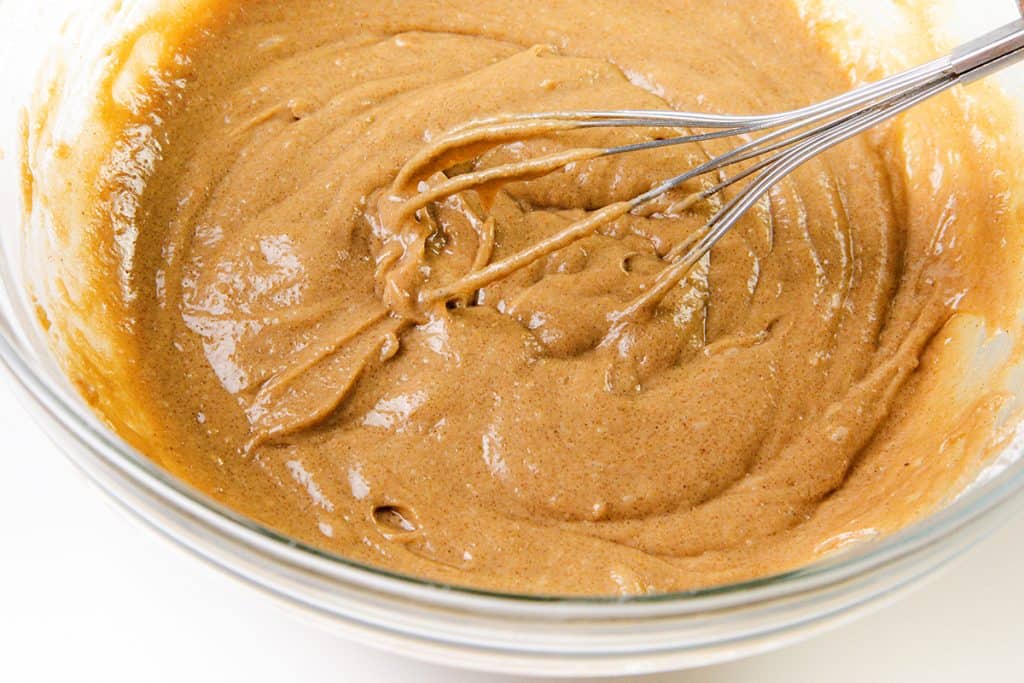 A metal whisk mixing thick, light brown gingerbread batter in a clear glass bowl on a white surface.