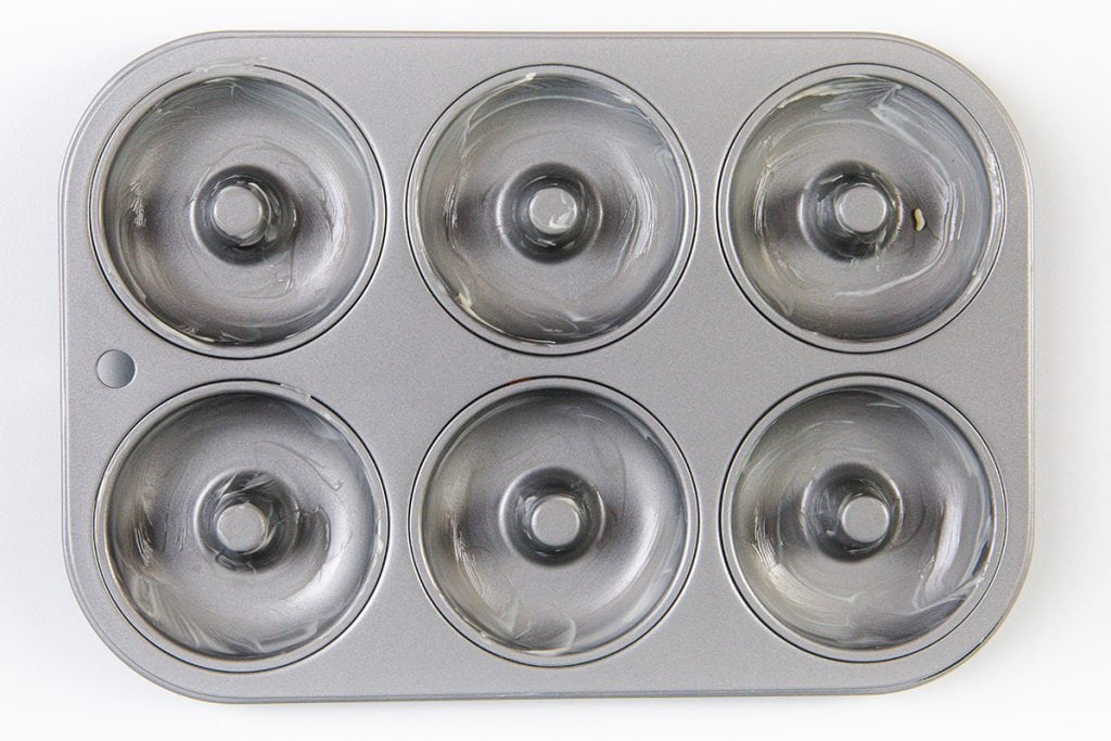 A six-cavity metal donut baking pan, each cavity coated in a thin layer of butter or oil—perfect for baking gingerbread or Christmas donuts, viewed from above.