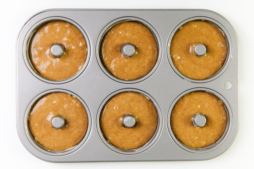 A metal donut pan filled with six cavities of unbaked gingerbread donut batter, ready to be baked, shown from above on a white surface.