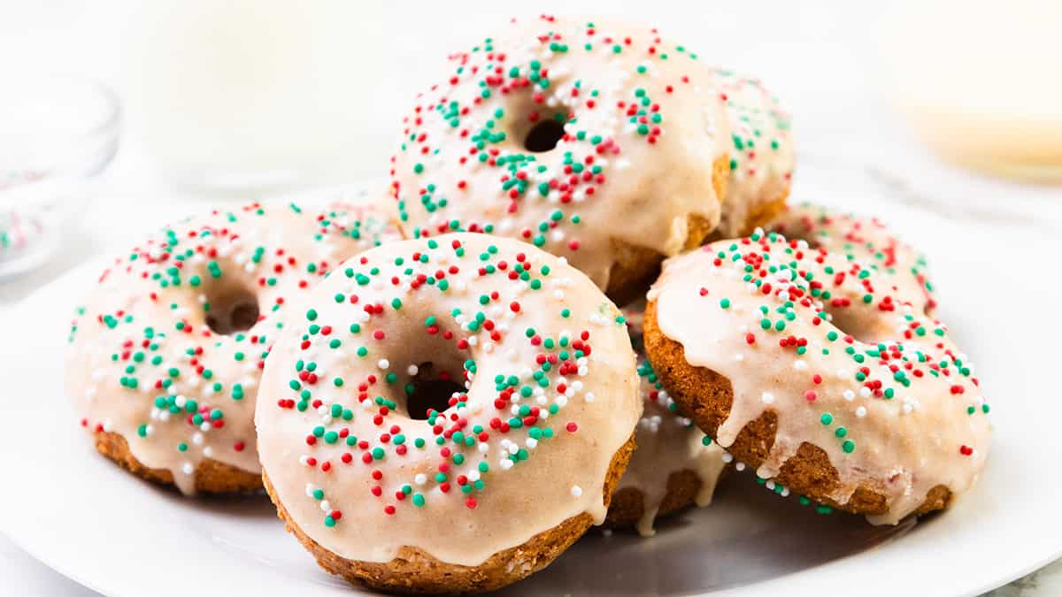 A plate of baked donuts topped with white icing and festive red, green, and white sprinkles, arranged in a pile on a white plate—perfect Christmas donuts for your holiday celebrations.