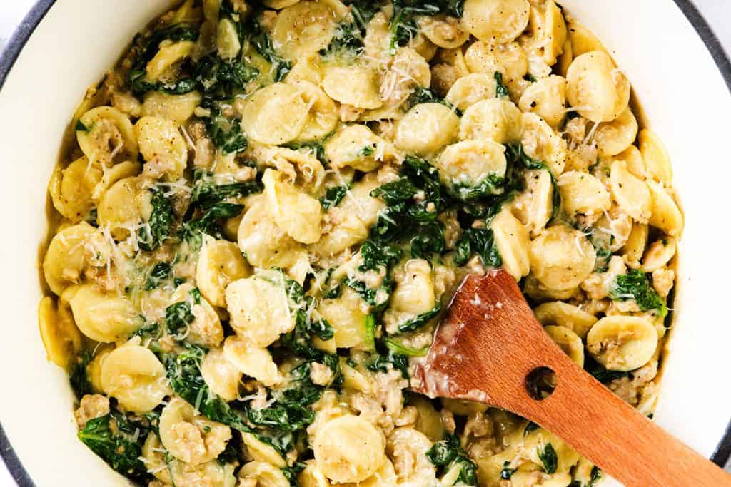 A close-up of creamy sausage orecchiette mixed with fresh spinach and grated cheese in a pot. A wooden spoon rests in this flavorful one-pot pasta dish, making it look irresistibly delicious.