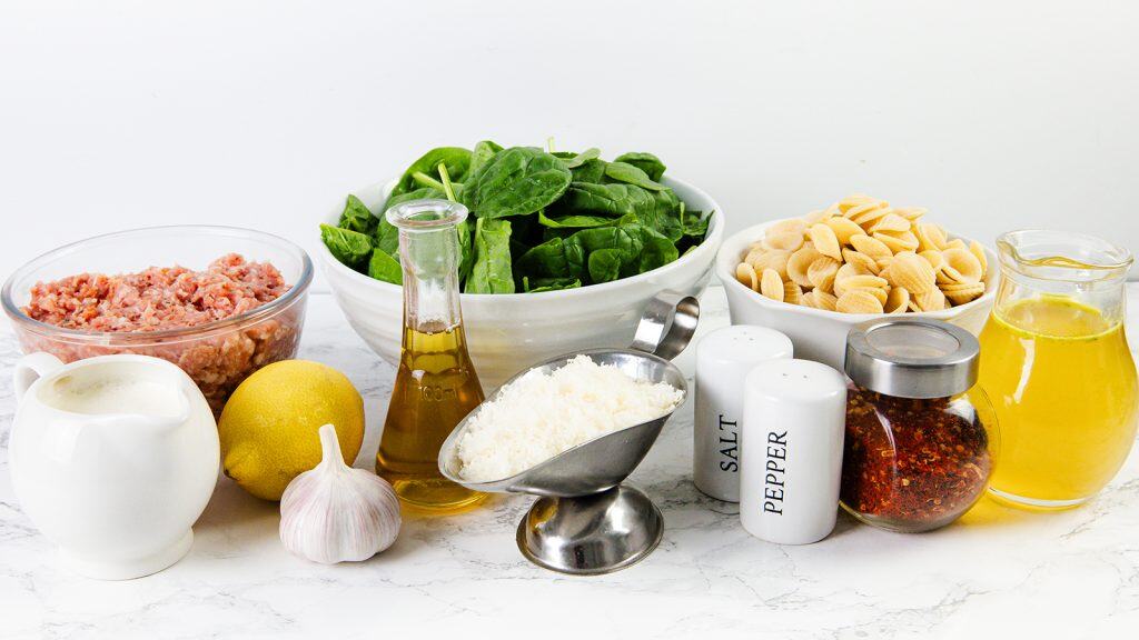 A variety of ingredients on a white surface, including ground meat for sausage orecchiette, fresh spinach, pasta shells, grated cheese, garlic, lemon, olive oil, cream, broth, salt, pepper, and chili flakes in jars and bowls.