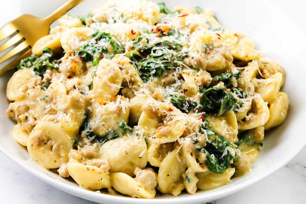 A white bowl filled with creamy sausage orecchiette pasta, spinach, and ground meat, topped with grated parmesan cheese and red pepper flakes. A gold fork rests on the side of this inviting one-pot recipe.