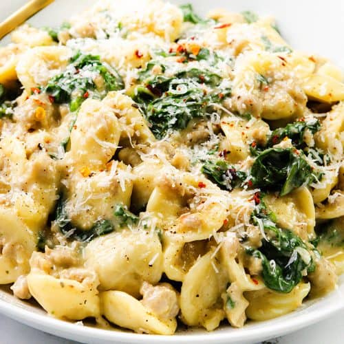 A white bowl filled with creamy sausage orecchiette pasta, spinach, and ground meat, topped with grated parmesan cheese and red pepper flakes. A gold fork rests on the side of this inviting one-pot recipe.