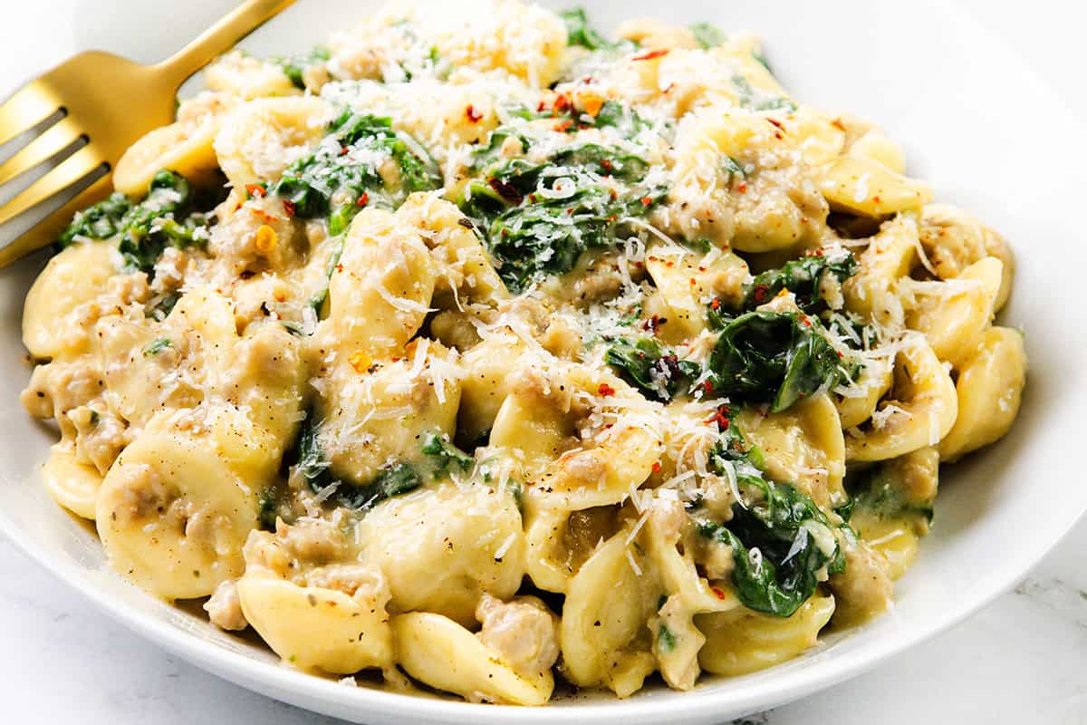 A white bowl filled with creamy sausage orecchiette pasta, spinach, and ground meat, topped with grated parmesan cheese and red pepper flakes. A gold fork rests on the side of this inviting one-pot recipe.