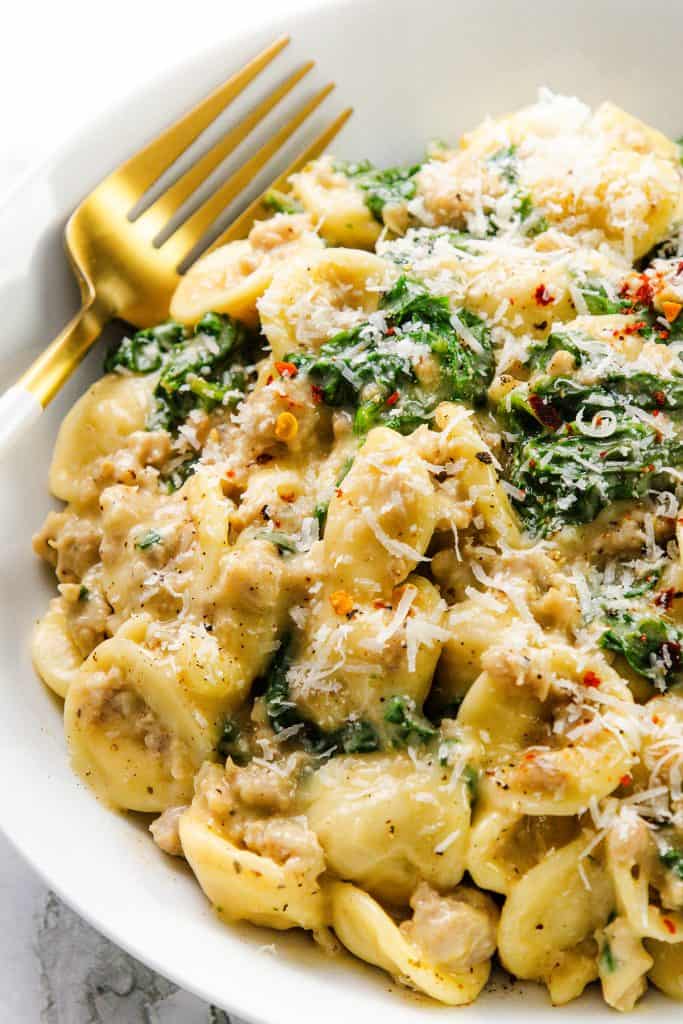 A close-up of a white bowl filled with creamy sausage orecchiette, spinach, and grated Parmesan cheese, with a gold fork resting on the side.