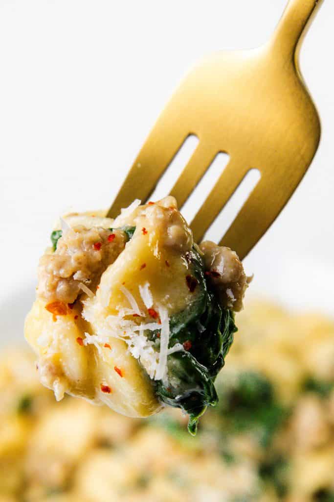 A close-up of a golden fork holding a bite of creamy sausage orecchiette with spinach, ground meat, grated cheese, and red pepper flakes, against a blurred bowl of pasta in the background.