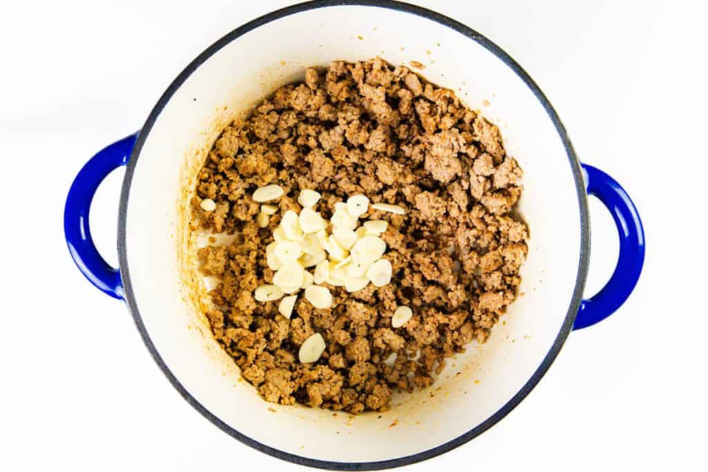 Cooked sausage in a white pot with blue handles, topped with sliced garlic cloves—perfect for starting a creamy sausage orecchiette—viewed from above on a white background.