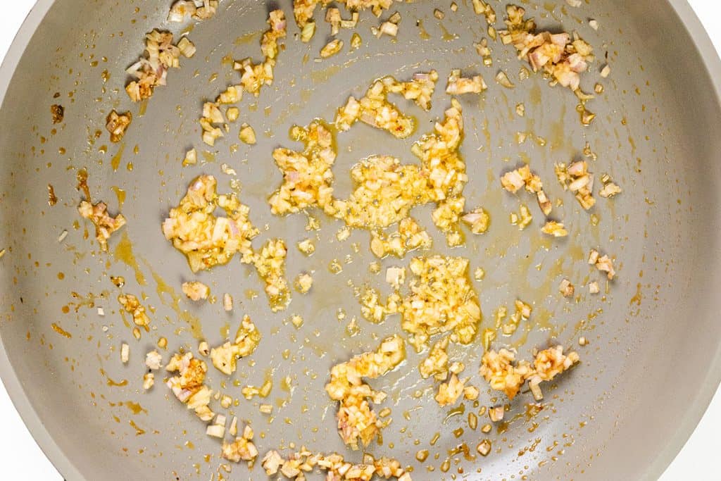 Finely chopped onions and garlic being sautéed in oil in a large light-colored frying pan, building the flavorful base for a one pot lemony shrimp orzo, with golden brown bits scattered across the surface.
