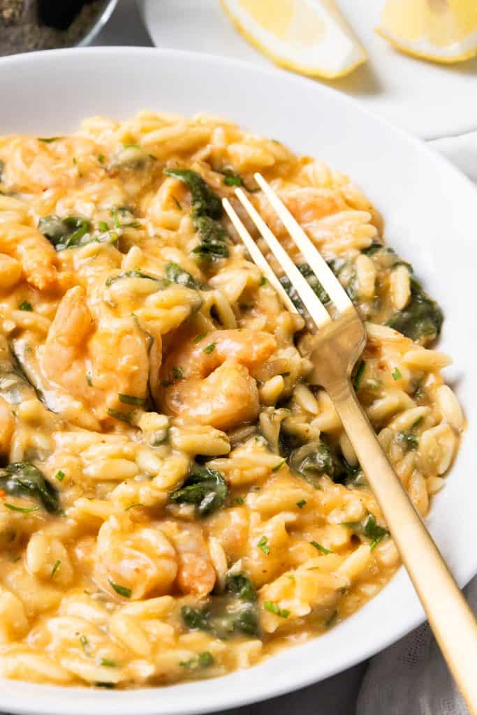 A white bowl filled with creamy lemony shrimp and spinach orzo, topped with herbs, with a gold fork resting on the side. This one pot dish is served with two lemon wedges in the background.