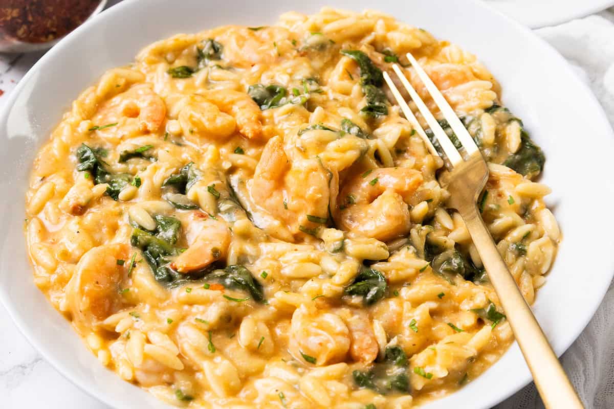 A white bowl filled with creamy orzo pasta mixed with lemony shrimp, spinach, and herbs, with a gold fork resting on the side.