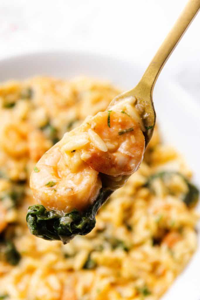 A close-up of a fork holding lemony shrimp, spinach, and creamy orzo above a plate filled with the same one pot dish. The food looks creamy and seasoned, with visible herbs adding a fresh touch.