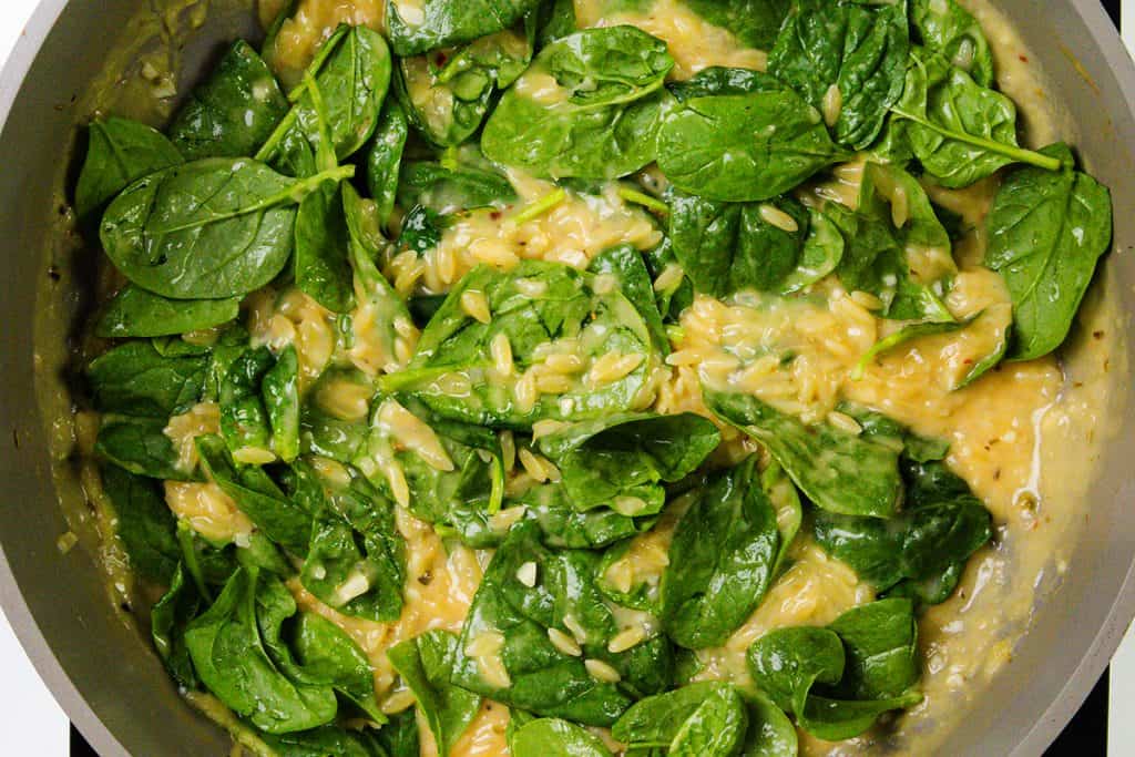 A close-up of a creamy one pot dish being cooked in a pan, featuring fresh spinach leaves mixed with orzo pasta in a thick, yellowish sauce.