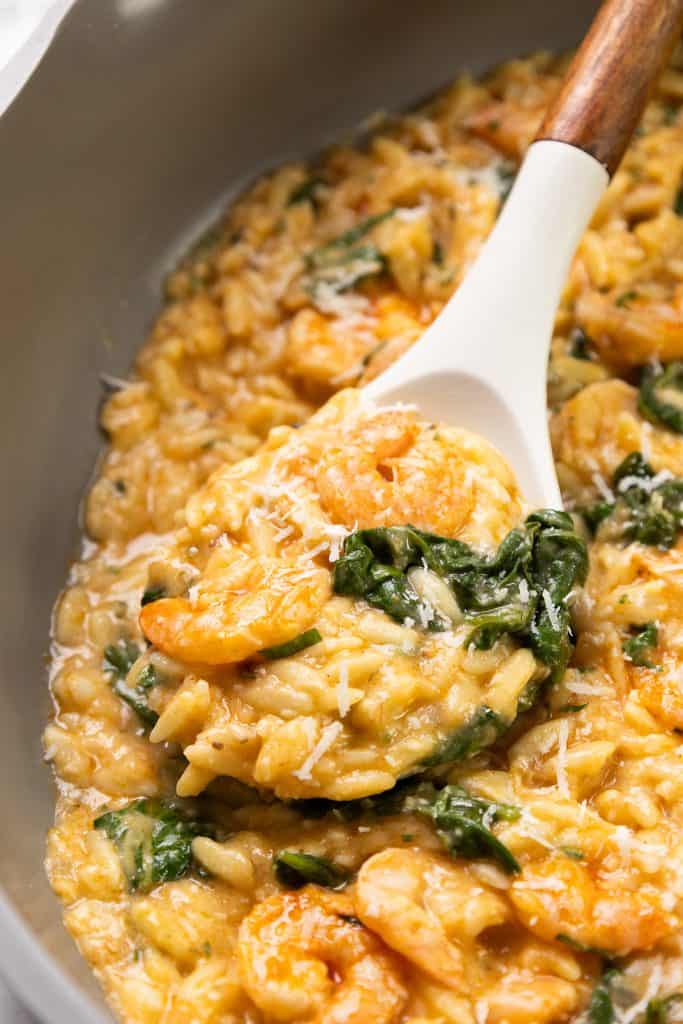 A close-up of creamy lemony shrimp and spinach risotto in a pot, with a white spoon lifting a serving. The one pot dish is garnished with grated cheese and has a rich, golden color.