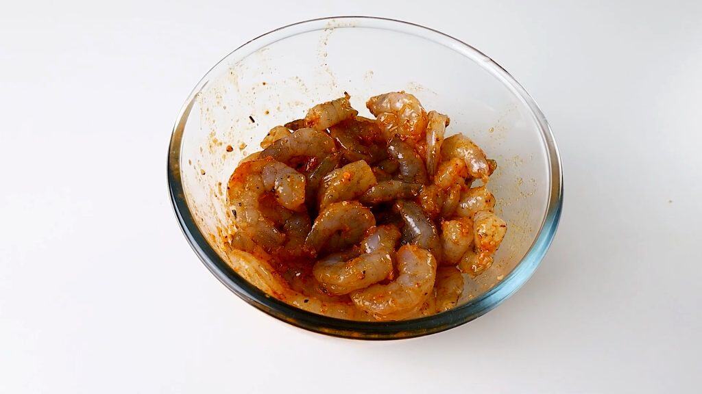 Raw shrimp coated in a reddish marinade with spices sit in a clear glass bowl on a white surface, ready to be transformed into a delicious one pot lemony shrimp dish.