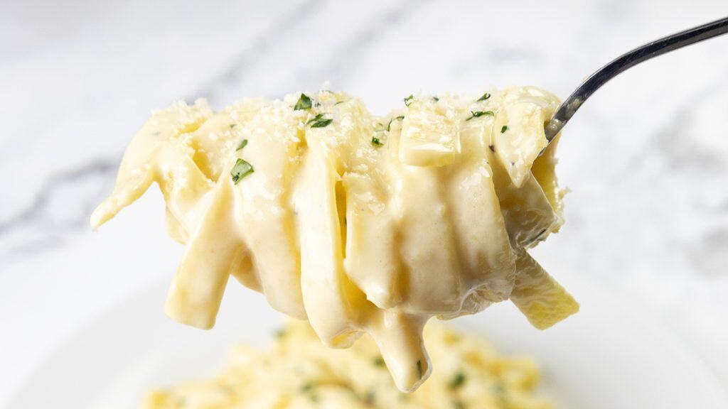 Tampilan jarak dekat dari garpu fettuccine krim Alfredo, menampilkan rasa pasta yang kaya dan dihiasi dengan peterseli cincang halus, dengan lebih banyak pasta terlihat di piring putih dengan latar belakang buram.