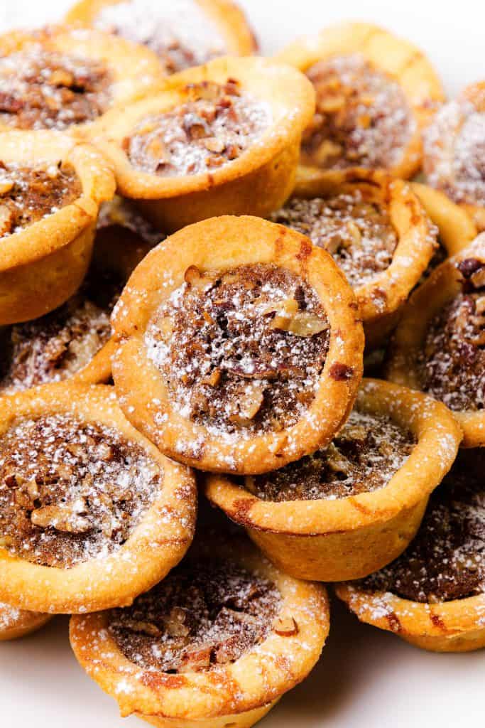 A close-up of a pile of Pecan Tassies dusted with powdered sugar, showcasing their golden-brown crusts and nutty filling.