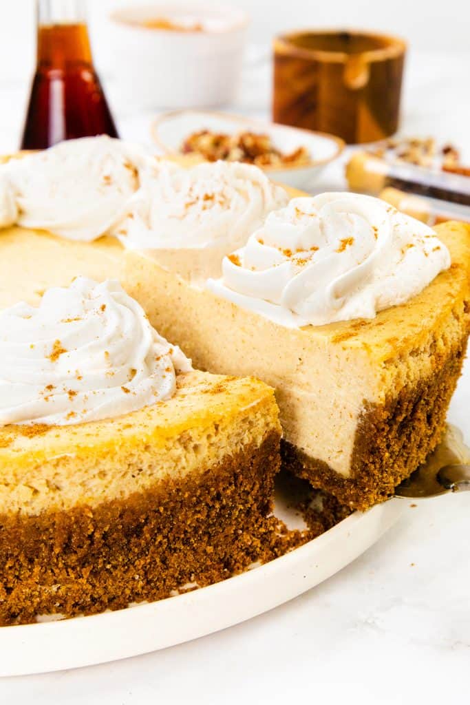 A creamy Pumpkin Cheesecake with a thick graham cracker crust is topped with swirls of whipped cream. One slice is being lifted out, revealing the smooth texture inside, while a few ingredients and dishes blur in the background.