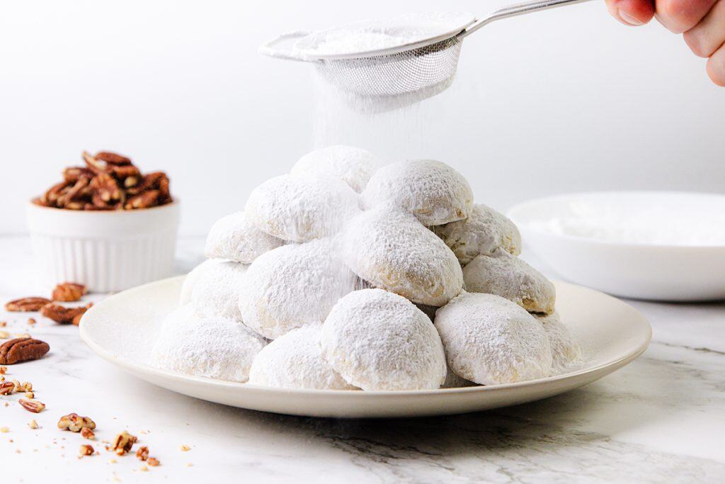 A hand sprinkles powdered sugar over a plate stacked with round Snowball Cookies. A bowl of pecans and a bowl of powdered sugar sit in the background on a white marble surface.