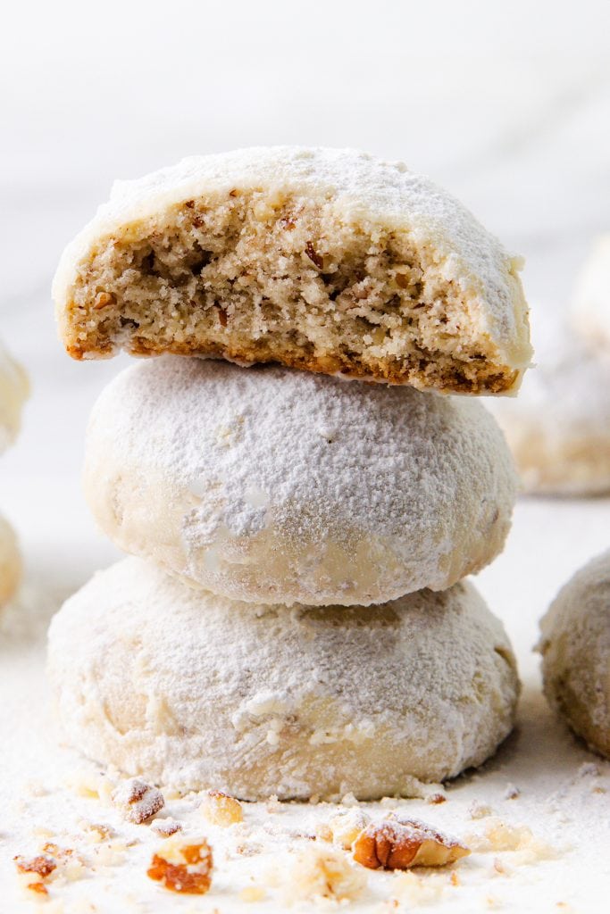 Three round Snowball Cookies coated in powdered sugar are stacked, with the top cookie showing a bite taken out, revealing a nutty, pecan-filled crumbly interior. Crumbs and chopped nuts are scattered in the foreground.