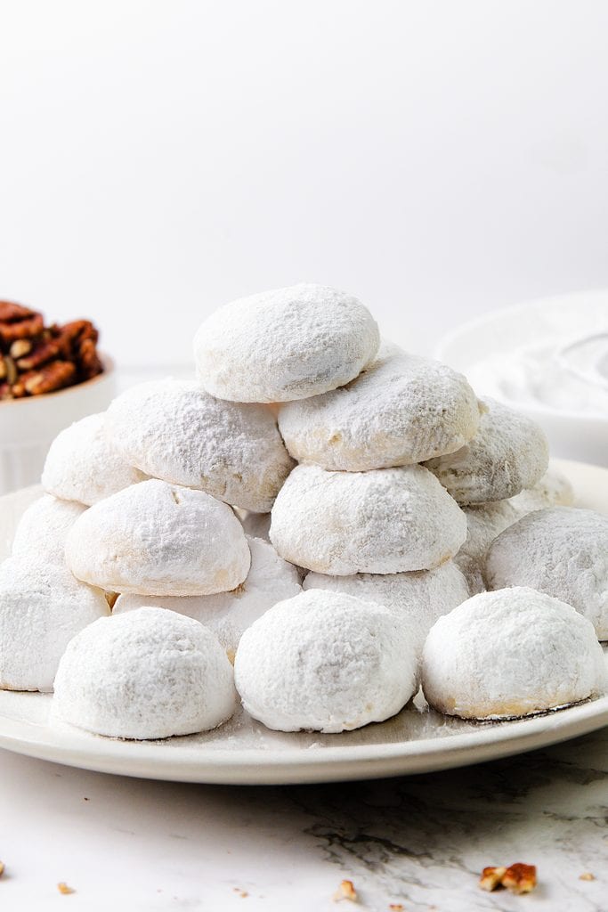 A plate stacked with round, powdered sugar-coated snowball cookies with pecans, arranged in a pyramid shape, with bowls of nuts and more cookies blurred in the background.