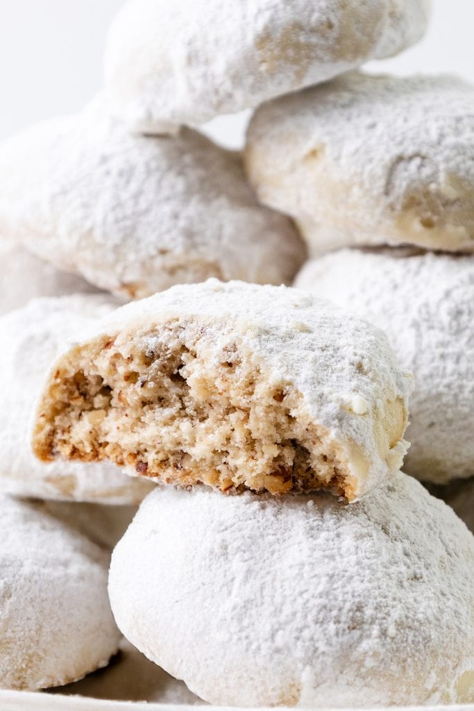A close-up of Snowball Cookies covered in powdered sugar, stacked on top of each other, with one cookie in the front partially bitten to reveal a soft, pecan-filled interior.