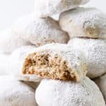 A close-up of pecan snowball cookies stacked in a pile, with one cookie at the front partially bitten to reveal a nutty, crumbly interior dusted with powdered sugar.