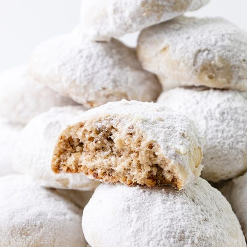 A close-up of pecan snowball cookies stacked in a pile, with one cookie at the front partially bitten to reveal a nutty, crumbly interior dusted with powdered sugar.