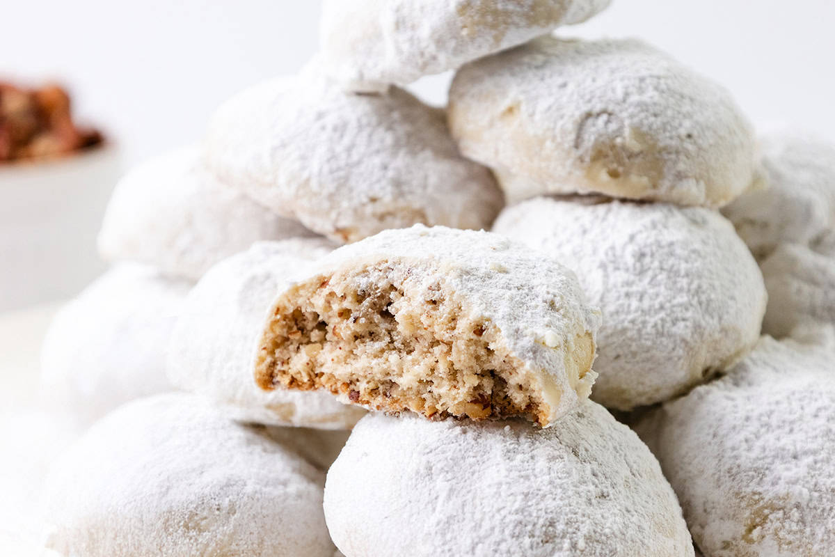 A close-up of pecan snowball cookies stacked in a pile, with one cookie at the front partially bitten to reveal a nutty, crumbly interior dusted with powdered sugar.