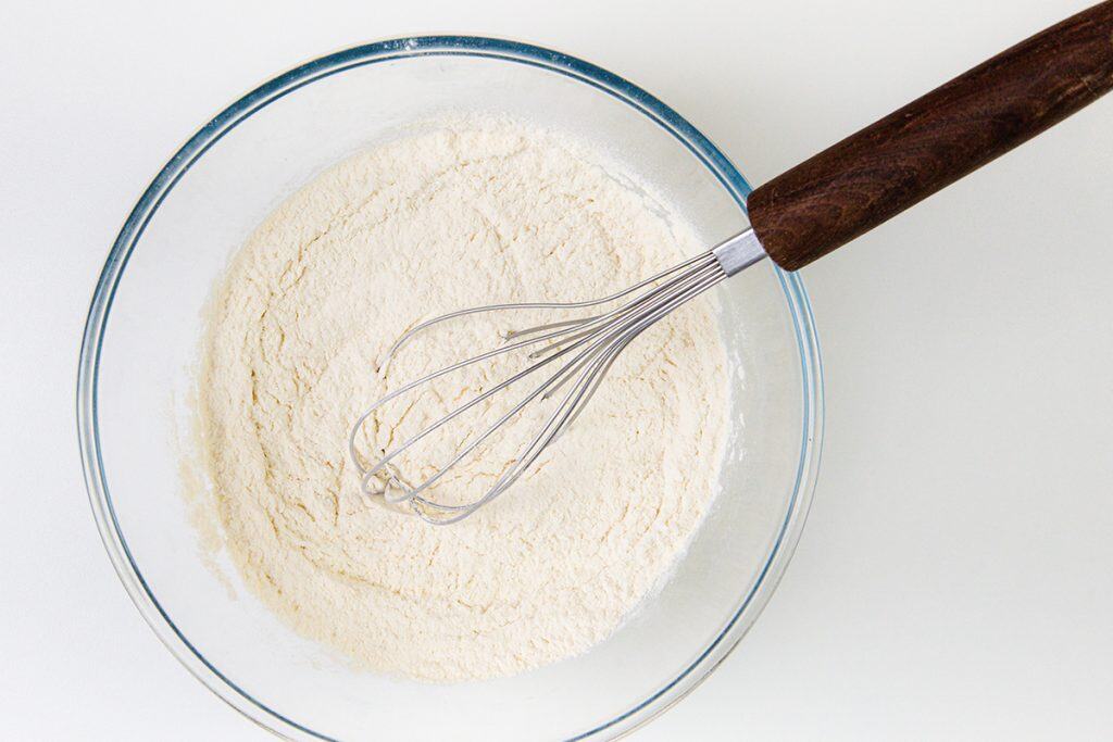 A glass bowl filled with flour and a metal whisk with a dark wooden handle resting inside, viewed from above on a white surface—perfect for mixing up dough for homemade Snowball cookies.